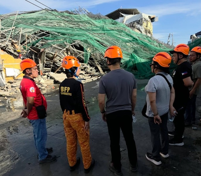 A group of people wearing protective hats stand in front of the collapsed building.