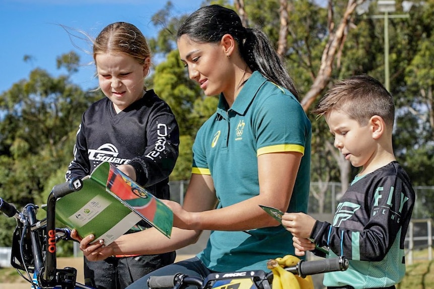 Saya Sakakibara sits on bike reading a magazine with two young kids next to her.