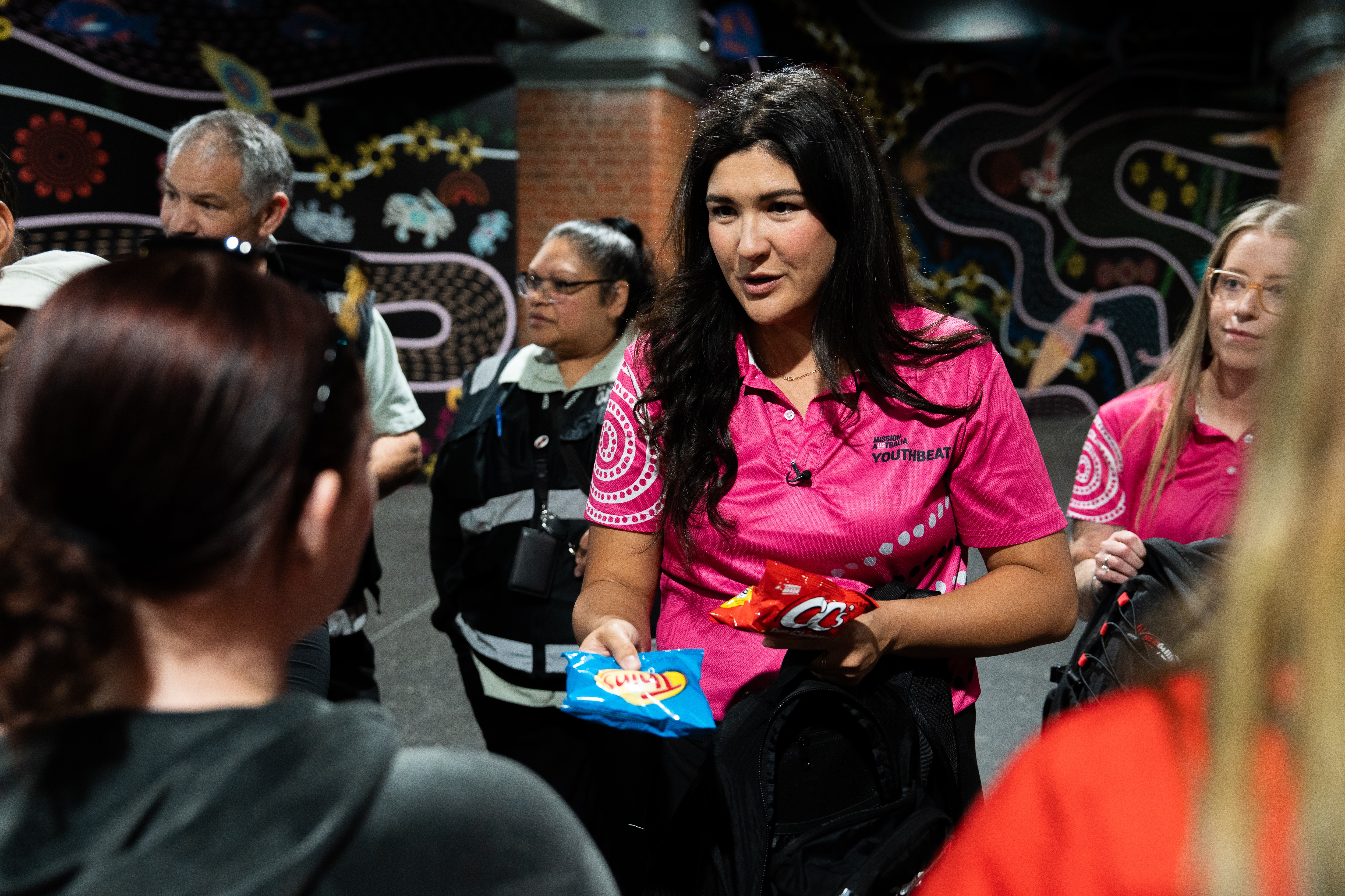 A close-up shot of a female youth worker with long black hair hands out a small packets of chips to a young girl.