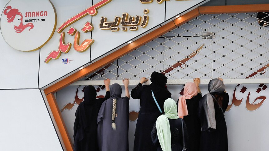 A group of women wearing burquas stand grabbing a beauty salon with orange writing 