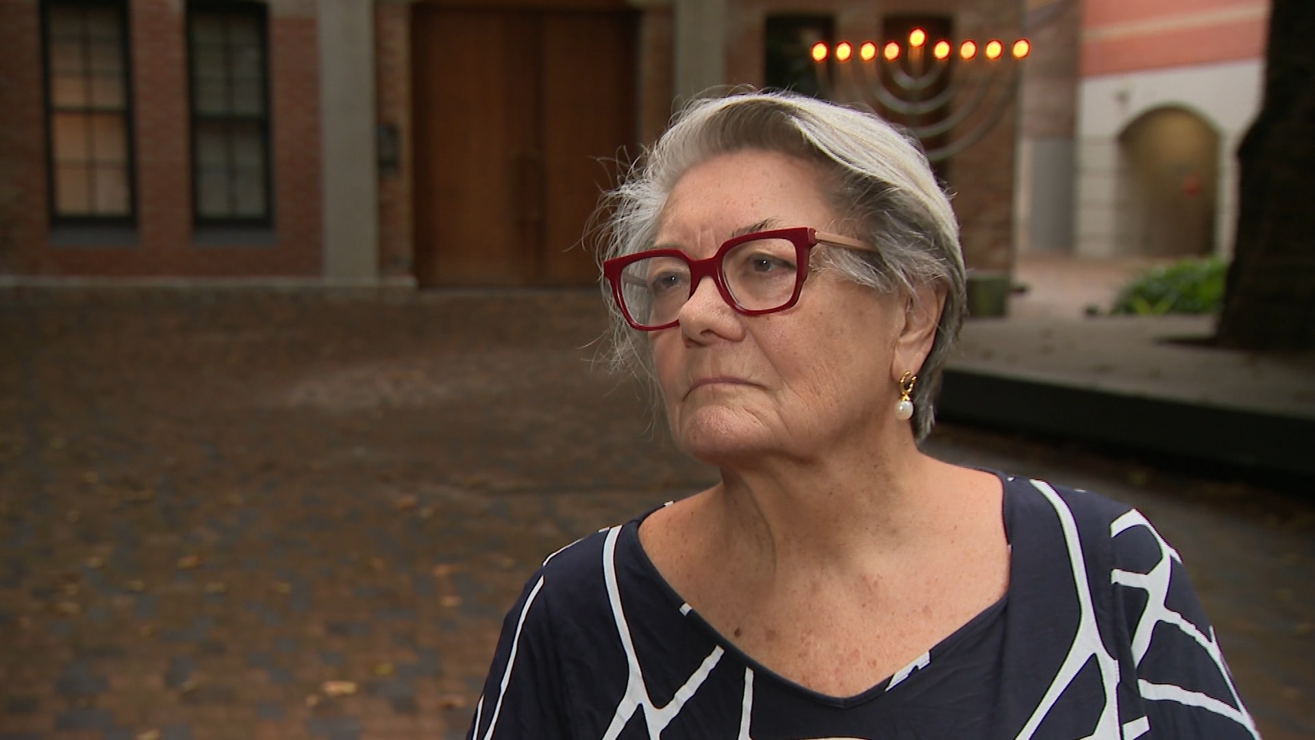 La Sra. Betts usa anteojos granates y está parada con el cabello recogido frente a un edificio de ladrillos y una menorá.