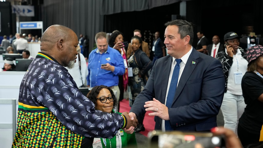 A middle-aged white man in a suit shakes hands with a middle-aged black man in a colourful shirt in a large hall.