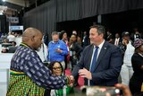A middle-aged white man in a suit shakes hands with a middle-aged black man in a colourful shirt in a large hall.
