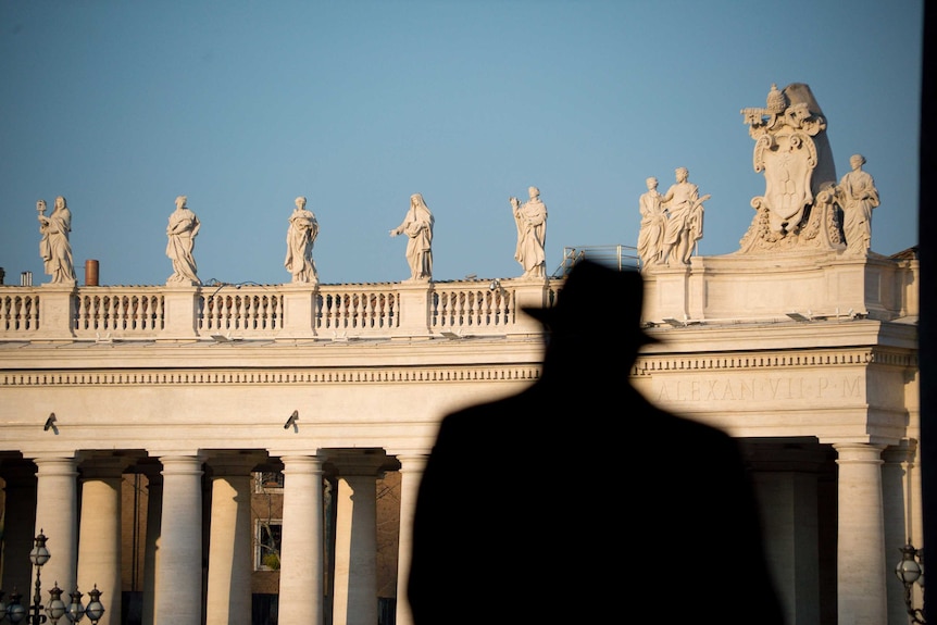 A man seen in silhouette walking away from the camera with statues in the background.