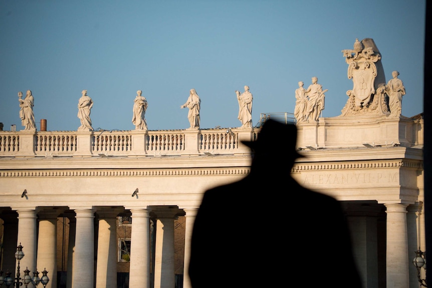 The shadowy outline of a man wearing a hat silhouetted against sunlit stone columns and statues