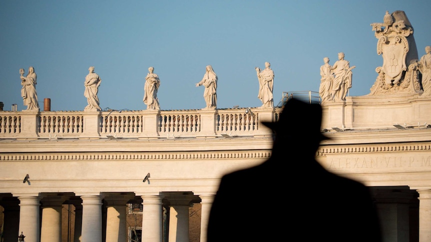 The shadowy outline of a man wearing a hat silhouetted against sunlit stone columns and statues