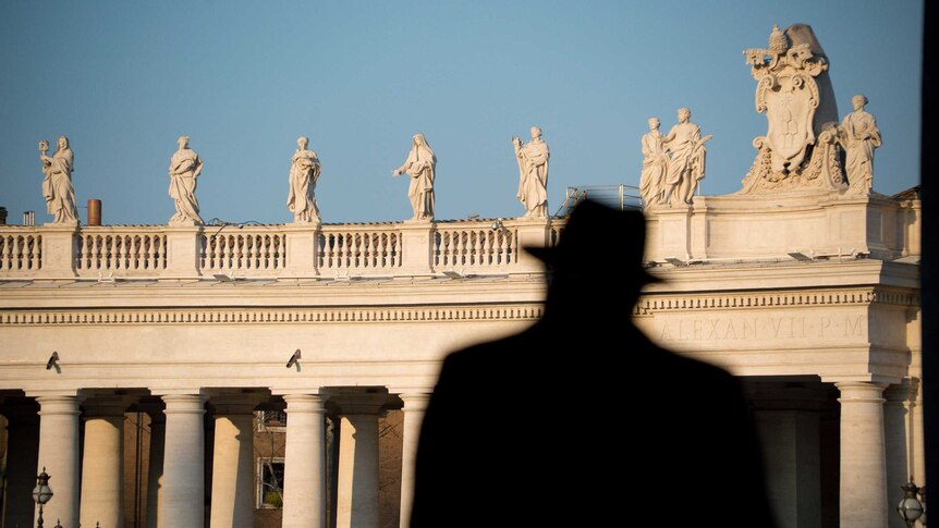A man seen in silhouette walking away from the camera with statues in the background.