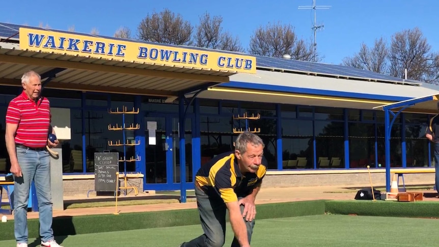 A man is bending over on a green surface with a lawn bowl rolling in front of him. There is a man watching from behind.