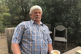 A man in a blue-checked shirt with grey hair and beard sits in a garden