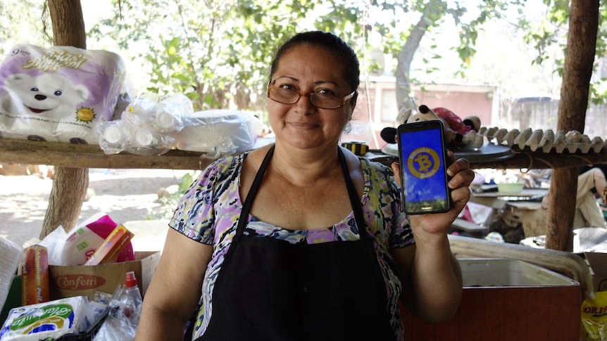 A woman in glasses and wearing an apron holds up her mobile phone showing the Bitcoin app
