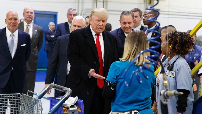 Donald Trump wearing a suit, meets two workers at a plant, machinery in the foreground, men in suits surround him.