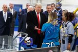 Donald Trump wearing a suit, meets two workers at a plant, machinery in the foreground, men in suits surround him.