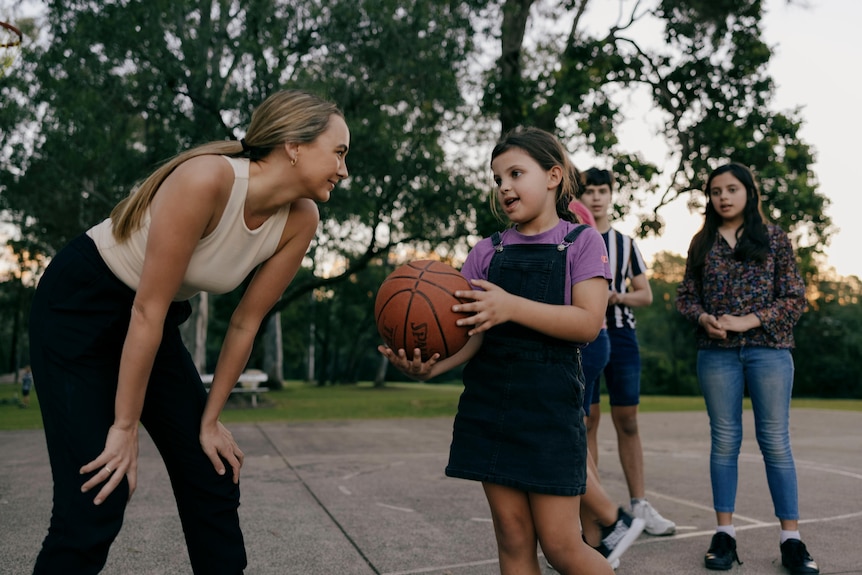 Young woman bending over, playing basketball with young girl in overalls and three other children at sunset