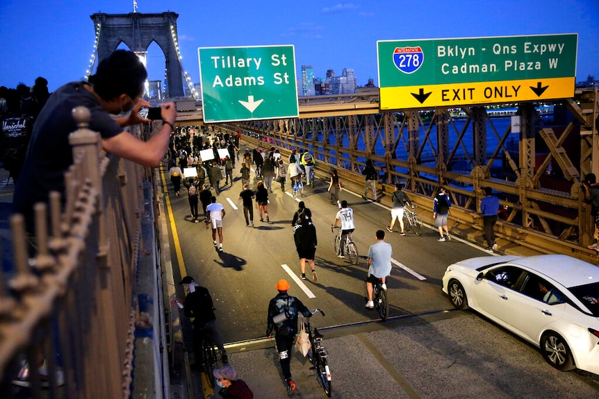From a high angle, you look down at the roadway of the Brooklyn Bridge filled with people walking across it at dusk.