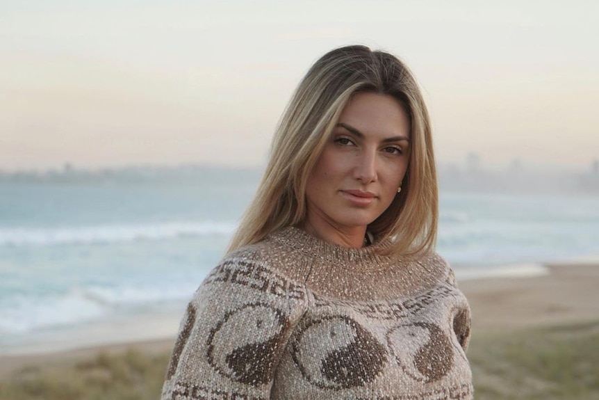 Oceana Strachan leans against a beach fence at dusk, wearing a brown jumper with the ocean behind her.