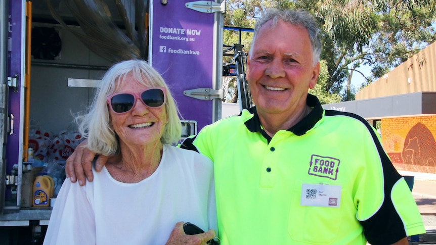 A man with a bright yellow Foodbank shirt puts his arm around a lady with sunglasses who is smiling