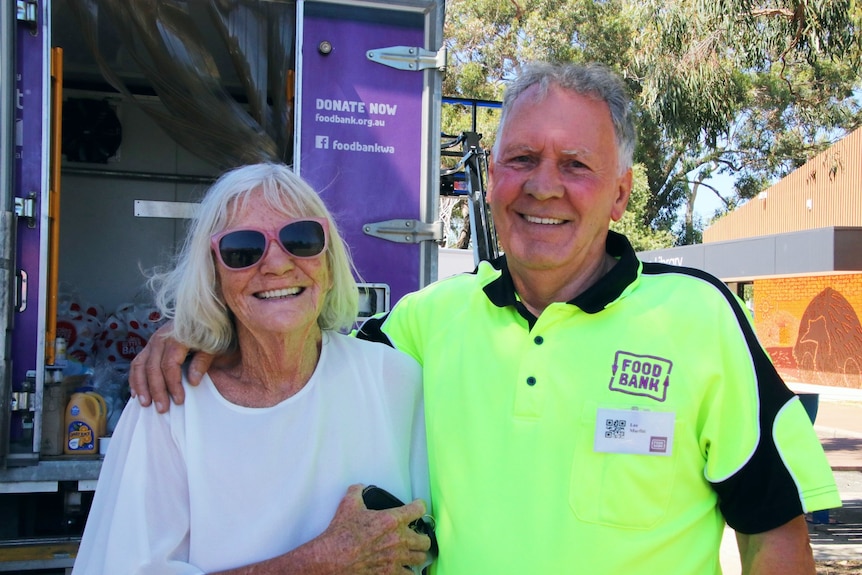 A man with a bright yellow Foodbank shirt puts his arm around a lady with sunglasses who is smiling