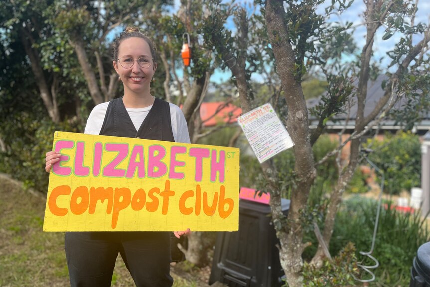 A woman holds a sign in front of a compost bin which reads "Elizabeth compost club." 