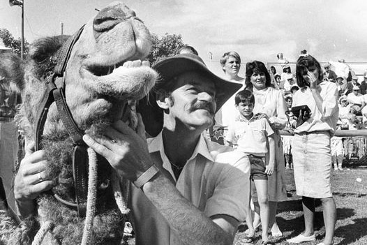 Black and white photo of Gordon O'Connell smiling widely as he holds a camel's face in his hands. Crowd of people stand behind.