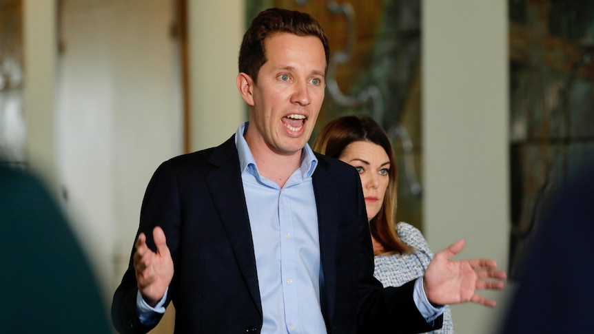 A man in a shirt and blazer with the top button undone gestures with his hands as he speaks.