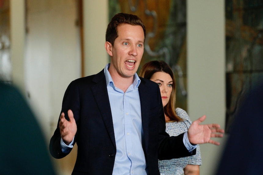 A man in a shirt and blazer with the top button undone gestures with his hands as he speaks.