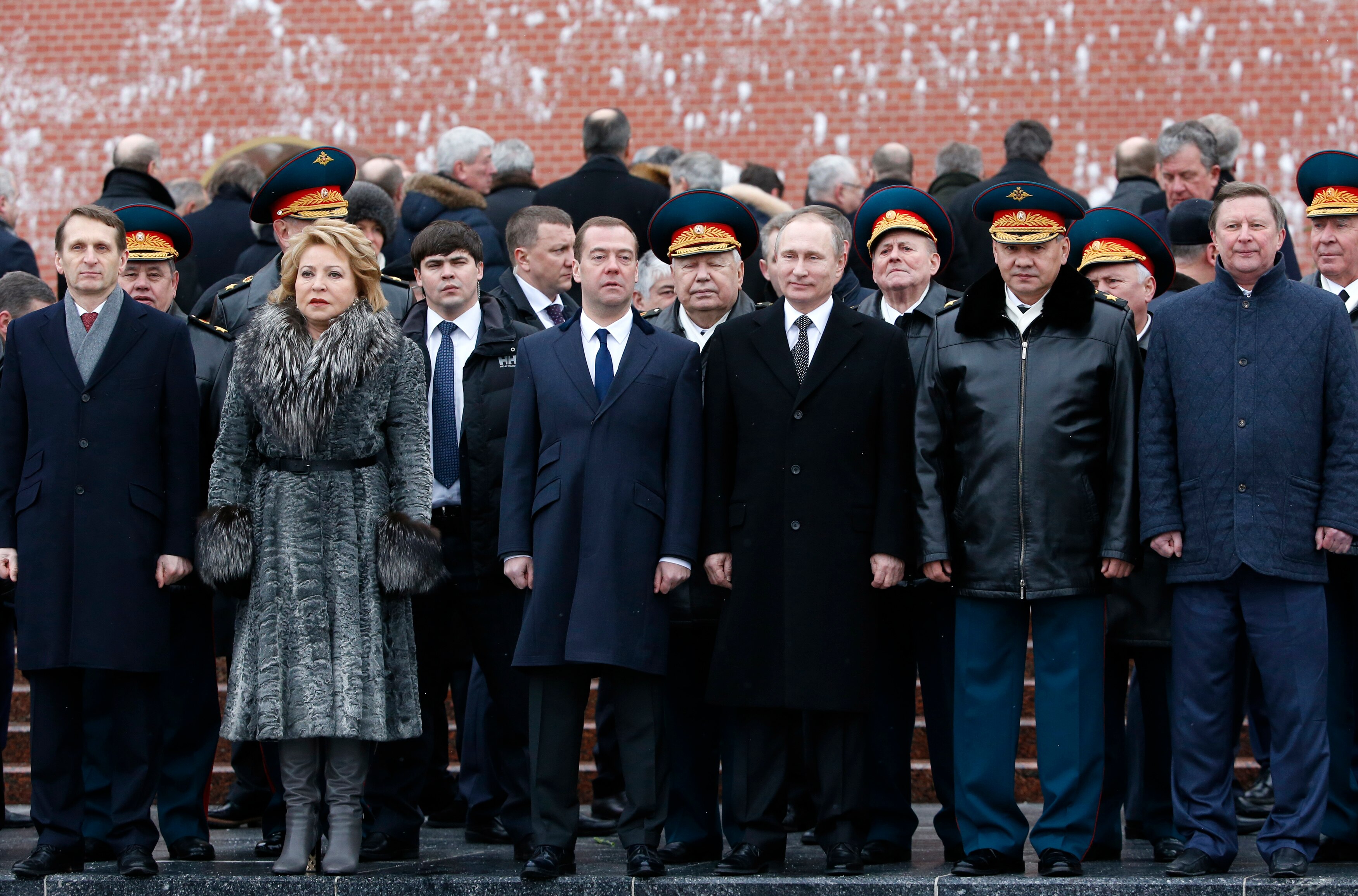 Vladimir Putin surrounded by a large group of men, and one woman with a blonde bob and dressed in a grey fur coat