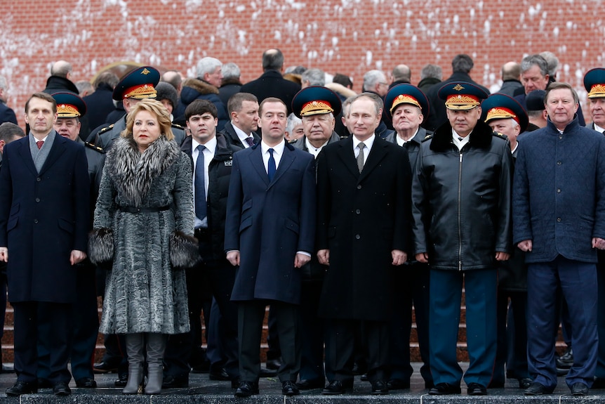 Vladimir Putin surrounded by a large group of men, and one woman with a blonde bob and dressed in a grey fur coat