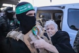 A masked man wearing a vest is supporting an elderly woman wearing a black coat