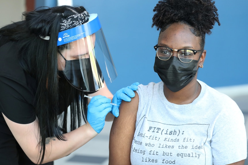 A dark-skinned woman with her curly hair piled high on her head receiving a vaccine from a nurse wearing limited PPE