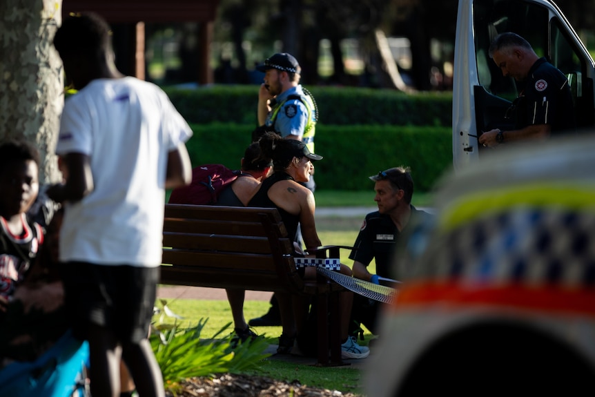 Two people sit on a park bench speaking to a paramedic from St John's WA in Burswood, with a police officer behind on his phone.