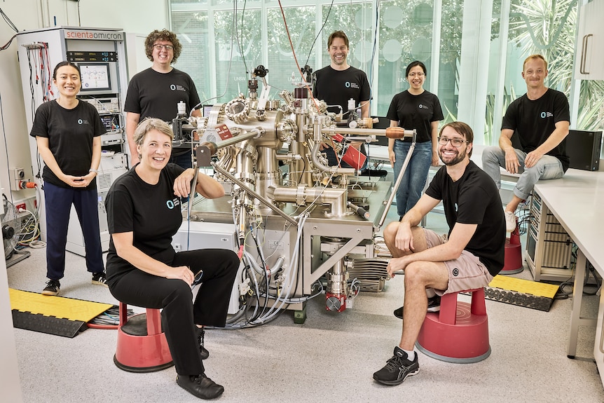 A group of seven people in dark t-shirts standing and sitting around a large piece of scientific equipment in a lab, smiling