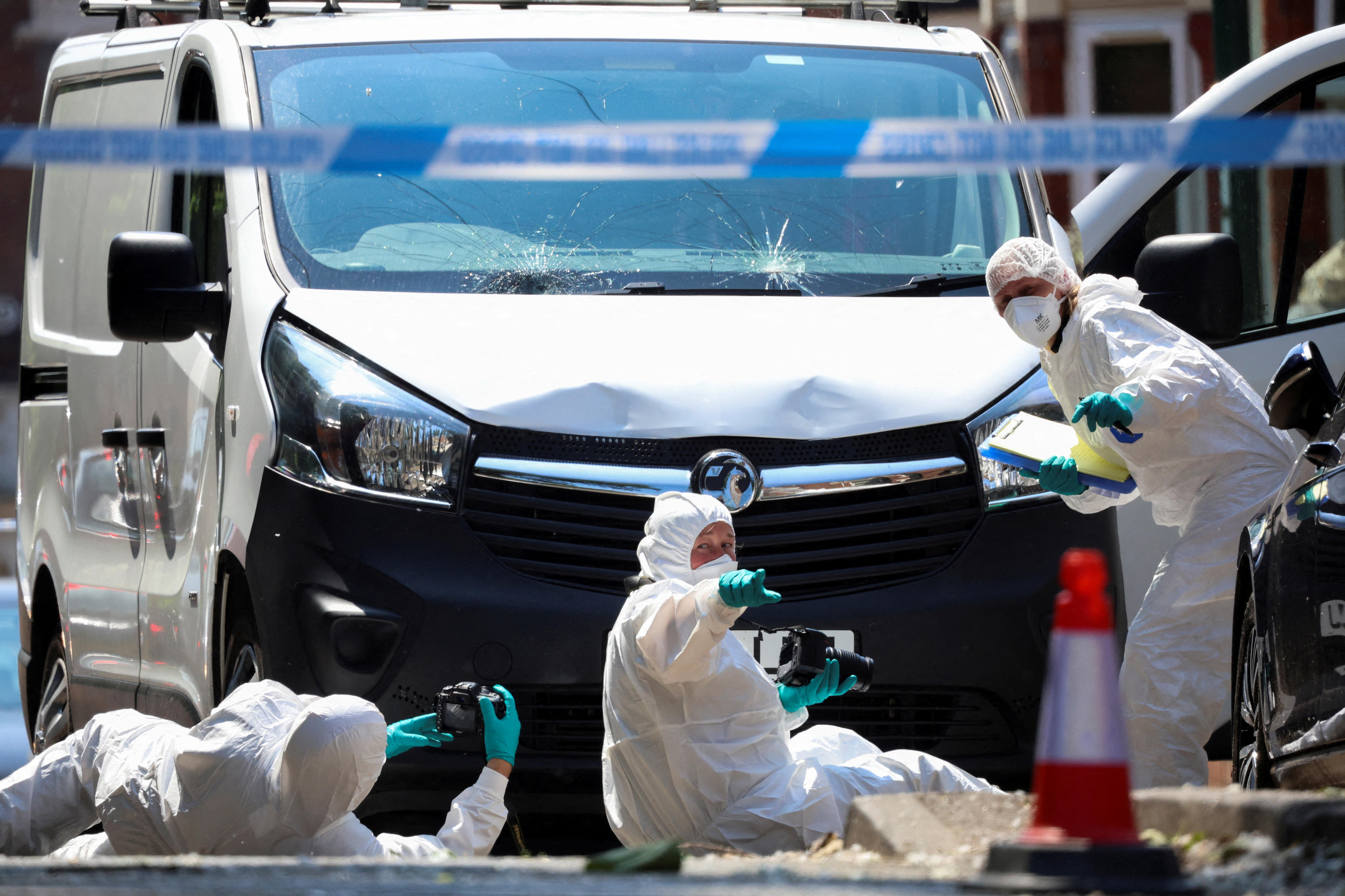 Three people wearing white forensic suits crouch at the front of a white van, which is dented with cracks in the windscreen