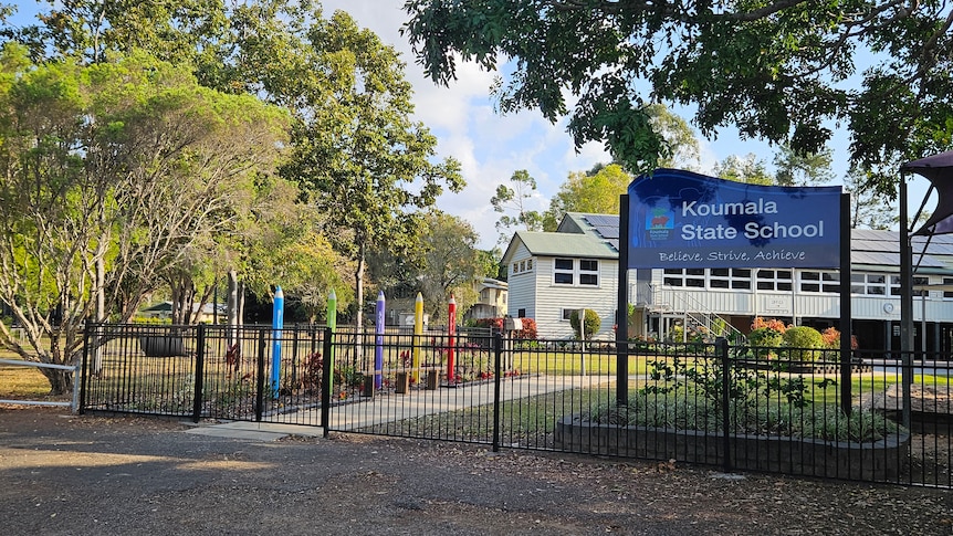 A sign that reads Koumala State School with a fence and buildings behind it, blue sky, green trees.
