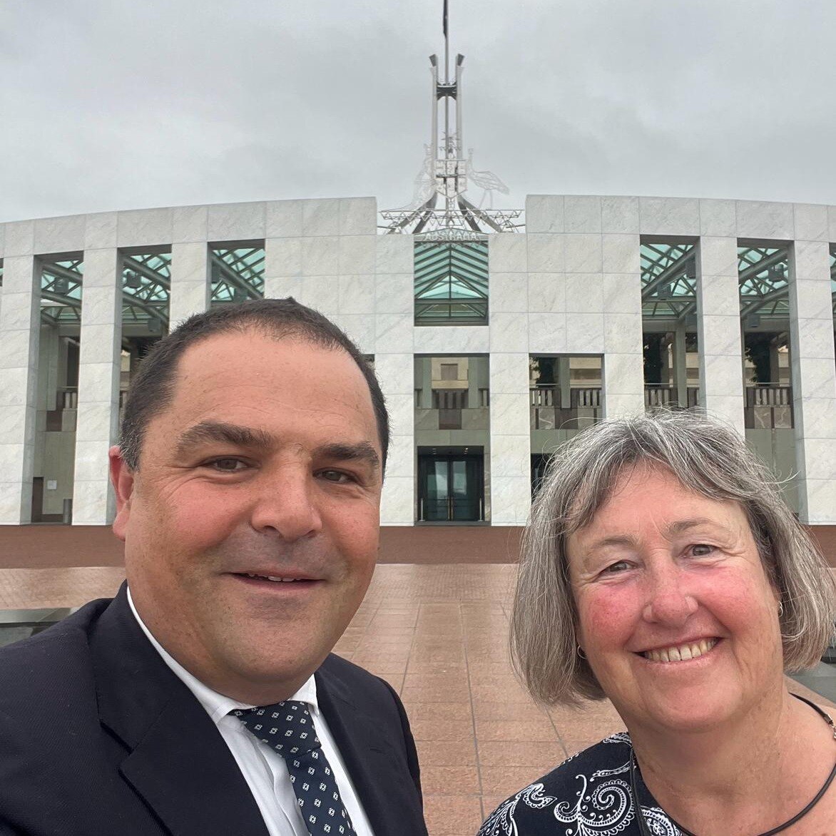 A man with short black hair with a woman with grey hair in front of Parliament House