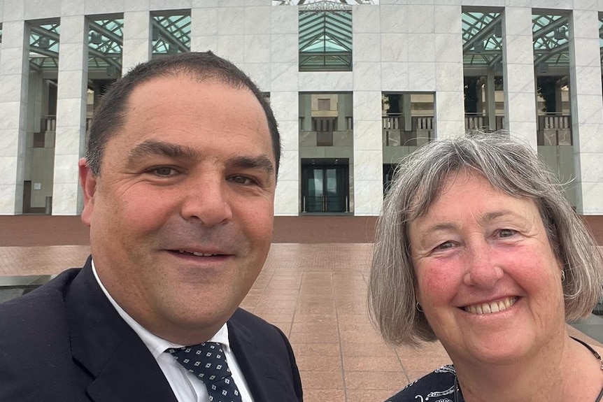 A man with short black hair with a woman with grey hair in front of Parliament House