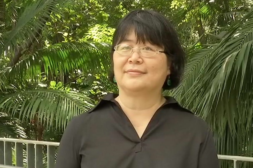 A woman with a fringe and short black hair, oval rimmed glasses and a black shirt with green earrings stands in front of ferns.