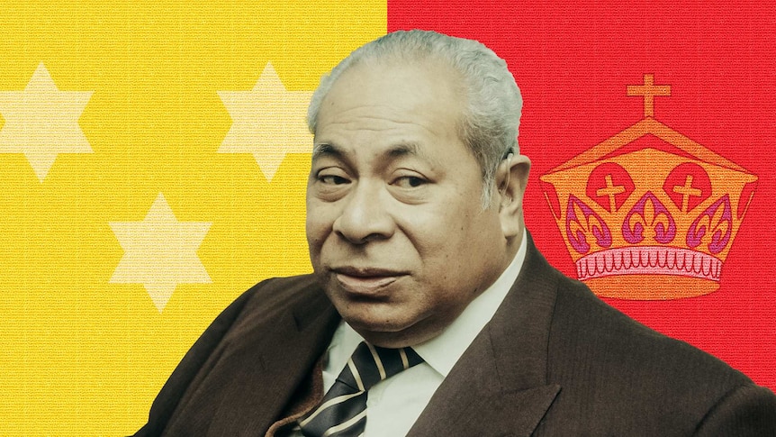 Tāufaʻāhau Tupou IV sits on a couch dressed in a suit. Parts of the Royal Standard of Tonga flag in background.