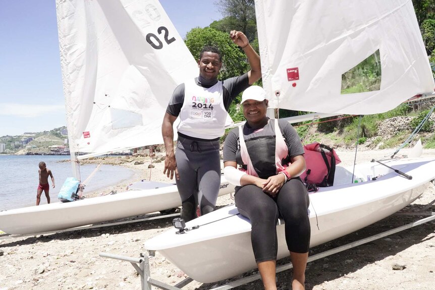 A man smiles as he stands leaning against a sail as a woman sits on a boat smiling.