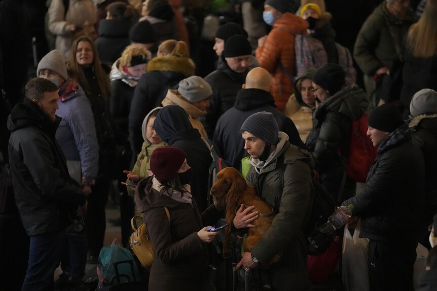a crowd of people in winter clothes wait at a train station where a man holds a dog next a woman holding a phone