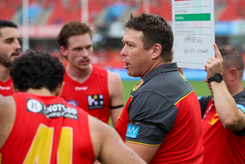 An AFL coach looks around his group of players speaking at quarter time, as the players look at a board.