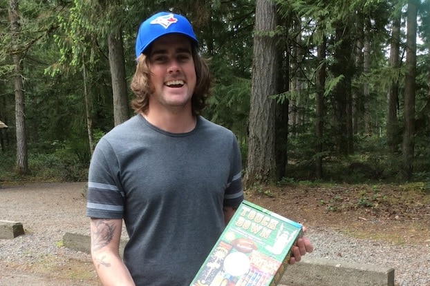 A man with brown hair smiling, he is wearing a blue hat and grey shirt and is in the woods