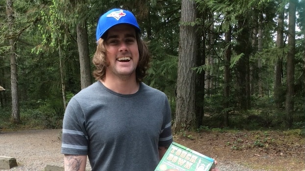 A man with brown hair smiling, he is wearing a blue hat and grey shirt and is in the woods.