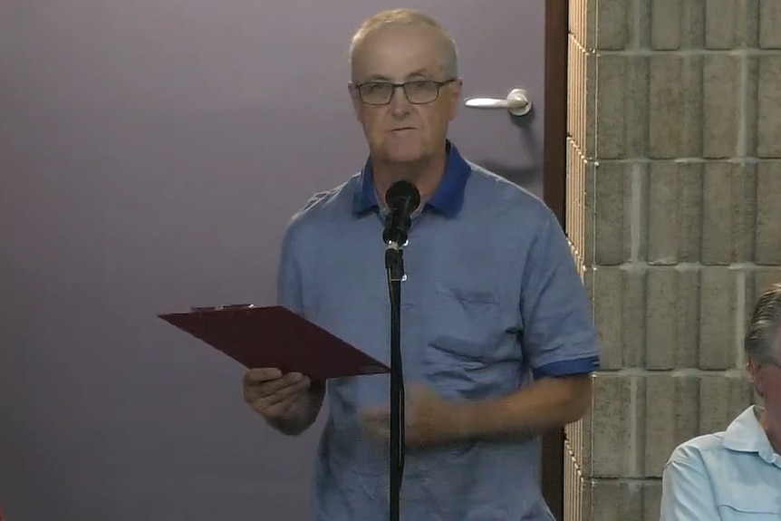 A man holding a clipboard speaks into a microphone.
