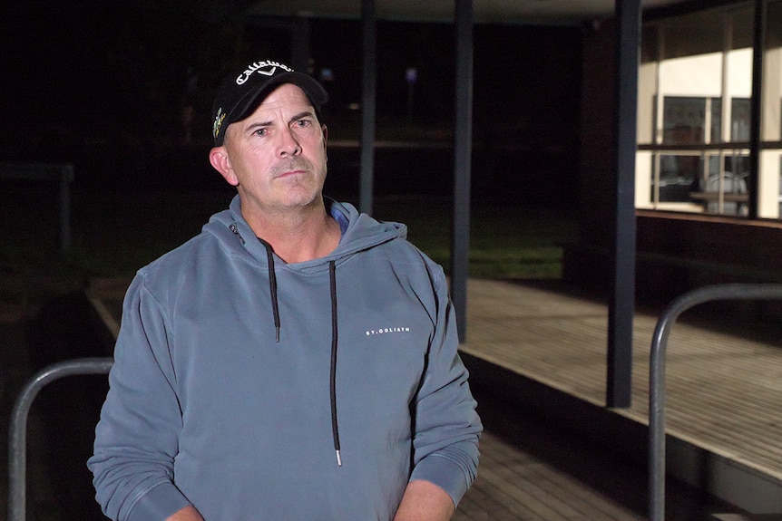 Brett wears a blue hoodie. He stands illuminated by a light outside the Orbost football club rooms.