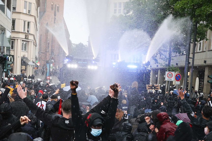 a crowd of people in the street raise their arms as a spray of water falls over them.