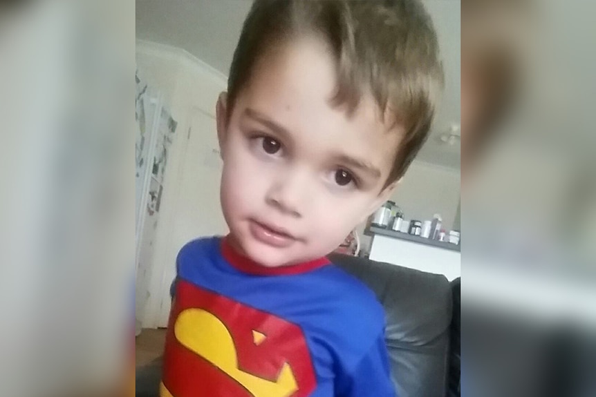 A close-up of two-year-old Lachlan Mitchell standing indoors wearing a Superman shirt.