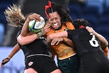 A Wallaroos player holds the ball while being tackled by two Canadian players.
