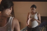 A pregnant indigenous woman stands in front of a mirror in a still from a video.