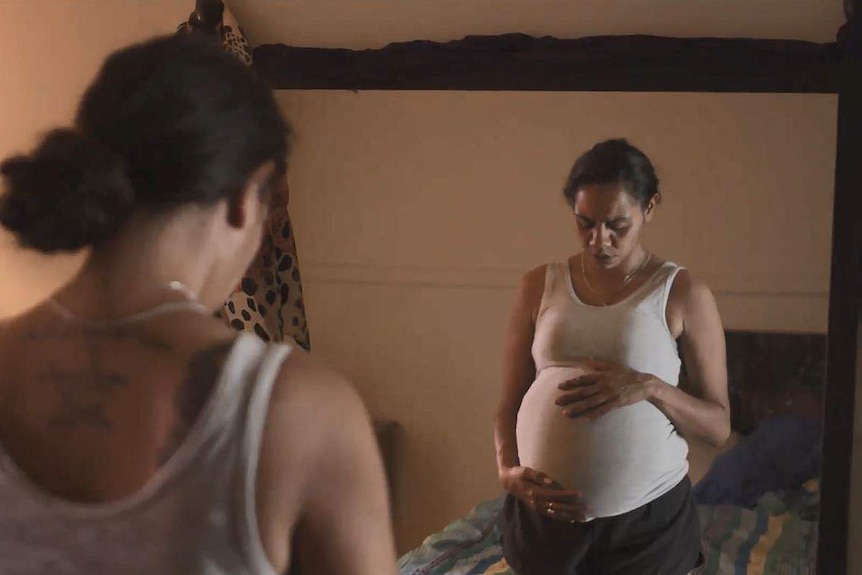 A pregnant indigenous woman stands in front of a mirror in a still from a video.