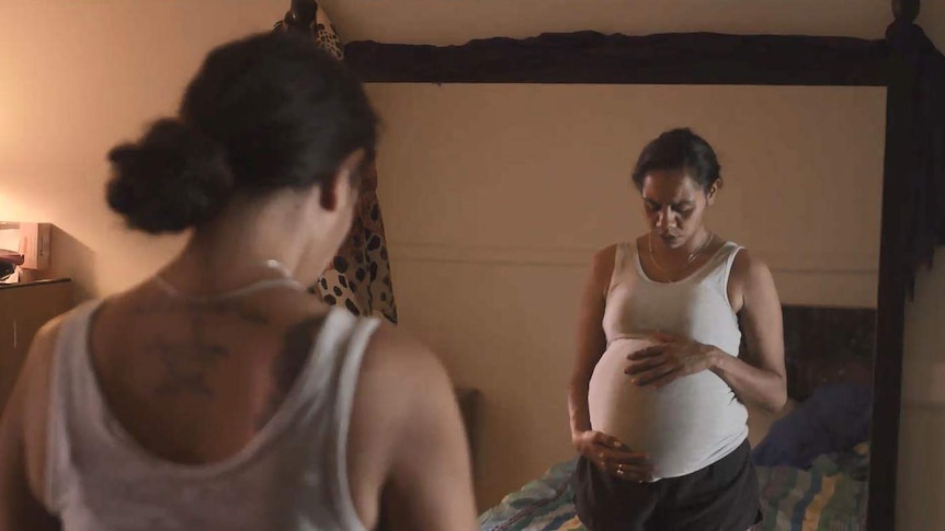 A pregnant indigenous woman stands in front of a mirror in a still from a video.